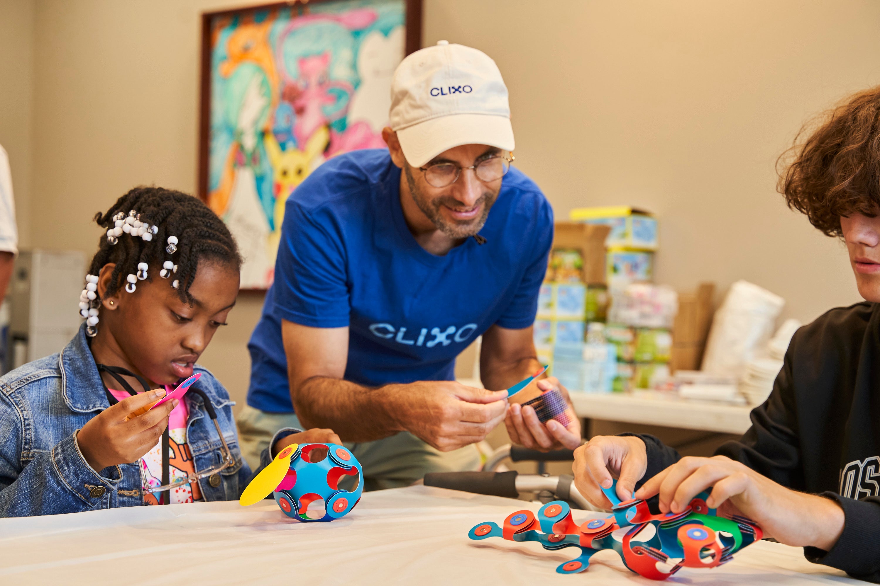 Assaf Eshet, a man wearing a blue T-shirt and tan baseball cap with "Clixo" written on them leans over a desk with two students seated - all engaged with building with Clixo pieces.