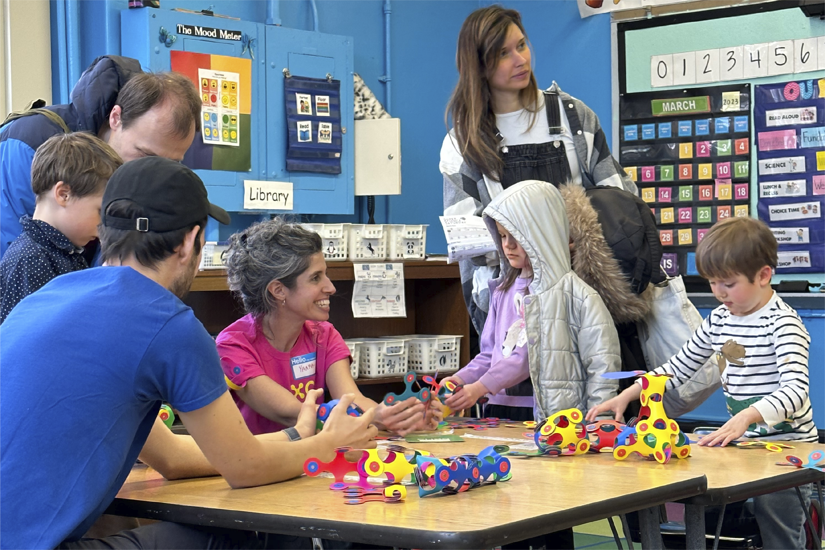 Two instructors wearing t-shirts with the word Clixo on them lean by a desk alongside multiple young students and parents building with Clixo pieces in a classroom.