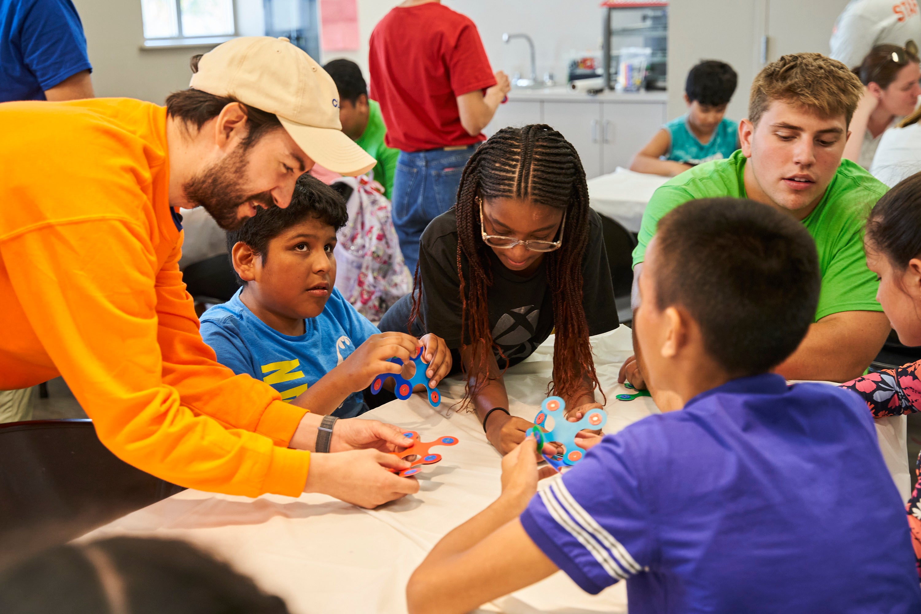 An instructor wearing a bright orange sweatshirt is leaning by a desk surrounded by students who are holding Clixo pieces. One student's eyes are gazing upwards.