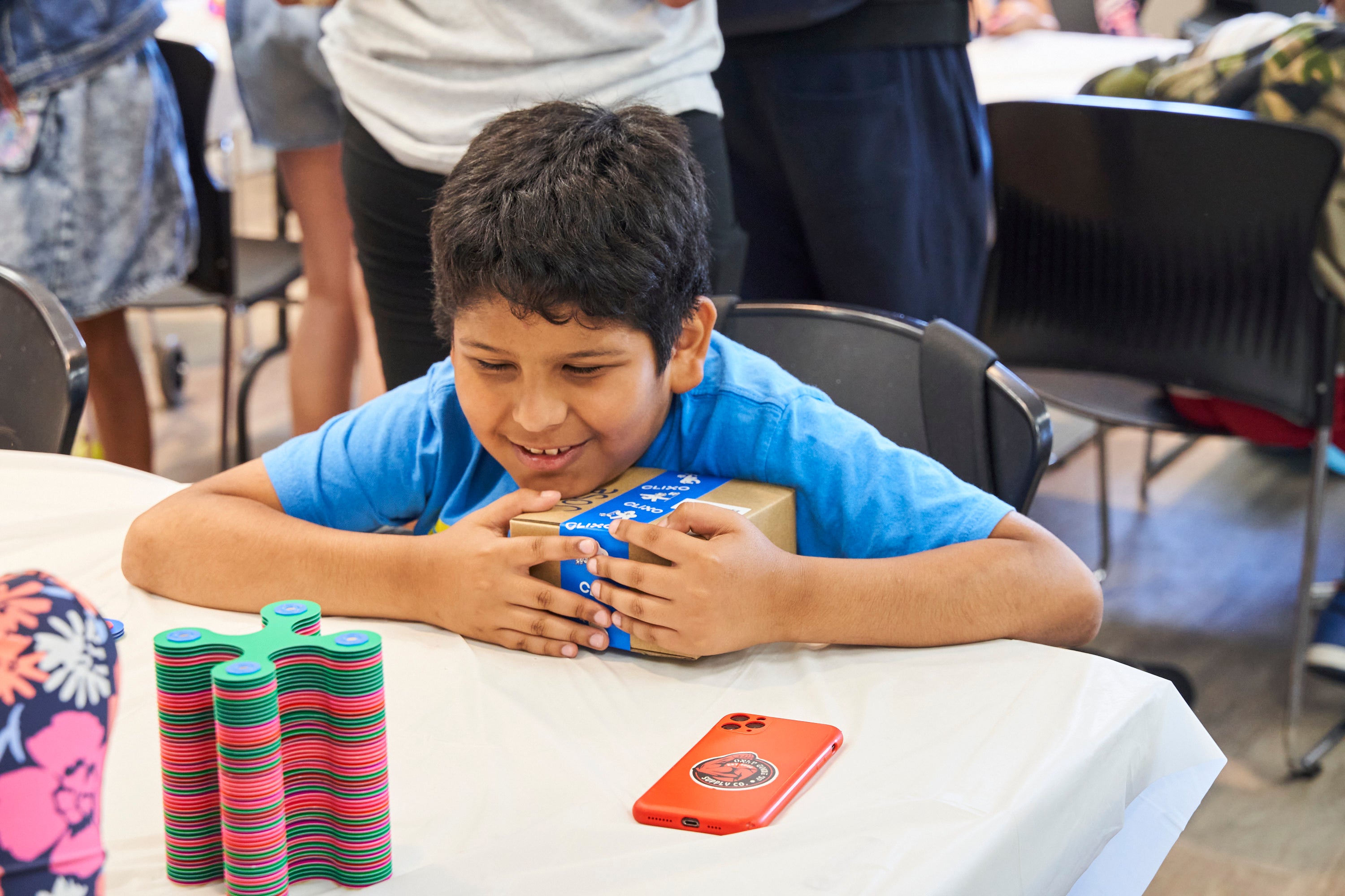 A young boy smiles while sitting by a desk hugging a cardboard box labeled Clixo. A stack of Clixo pieces in front of him.